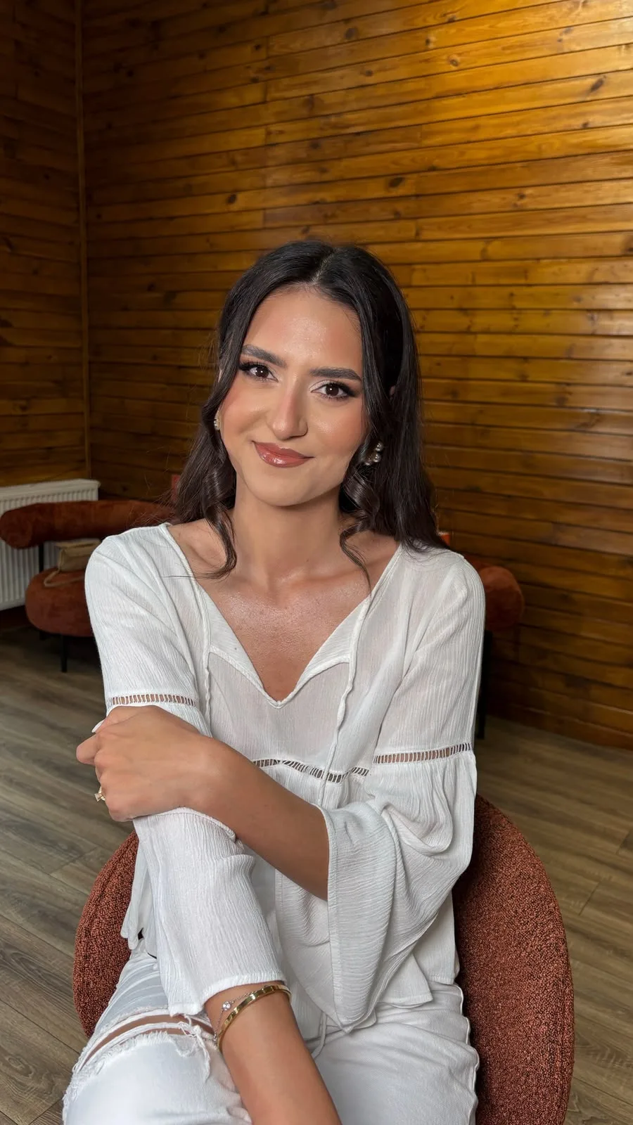 Clientă machiată în studio, cu bluză albă, în fața peretelui de lemn și lângă fotoliul terracotta